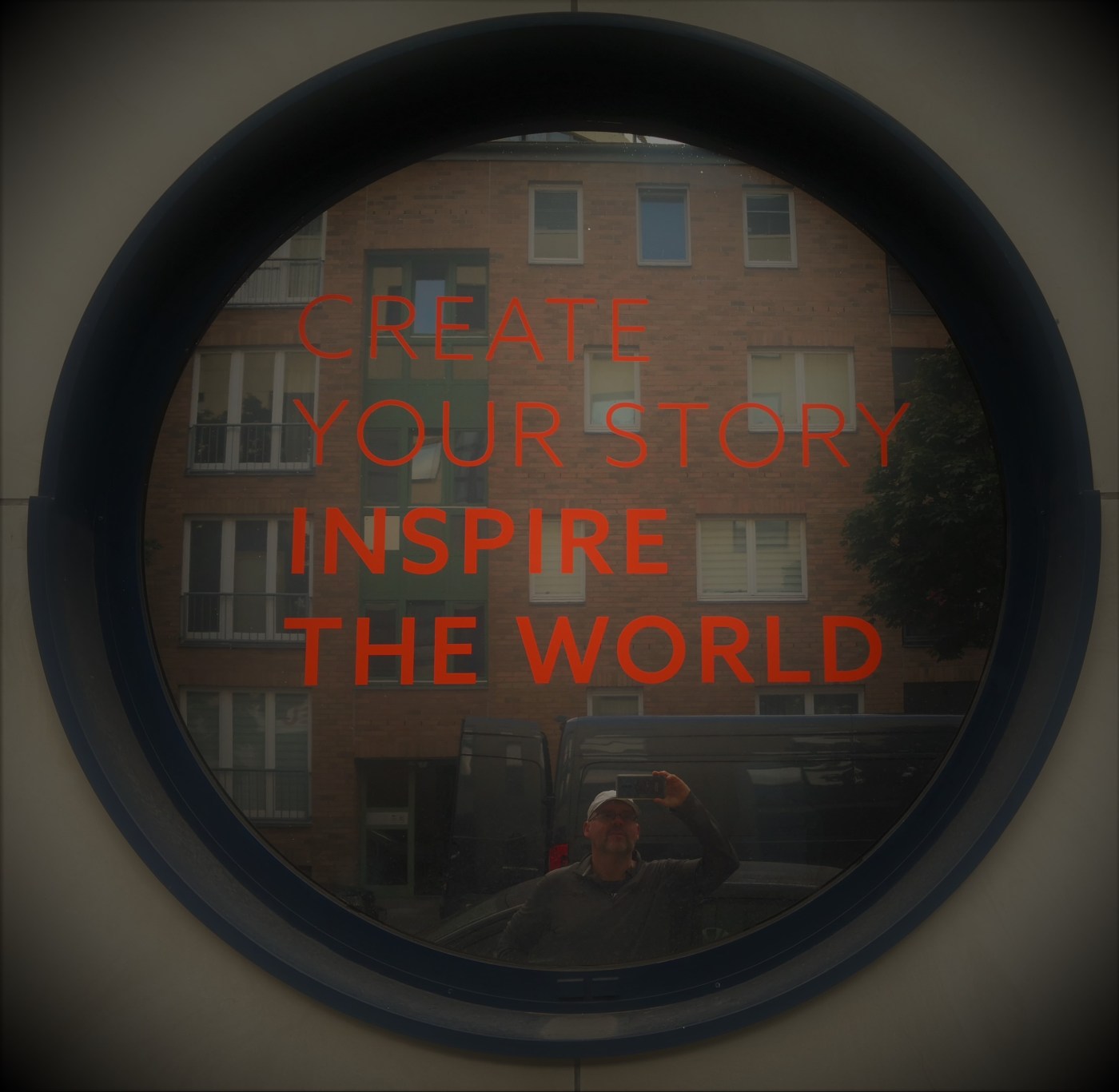 Bild zeigt ein Fenster in Bullaugen-Form, auf der man in roter Schrift lesen kann: "Create your Story, Inspire the World". In der Spiegelung der Fensterscheibe ist der Autor Ralph Raule schemenhaft zu erkennen, wie wer das Bild fotgrafiert.